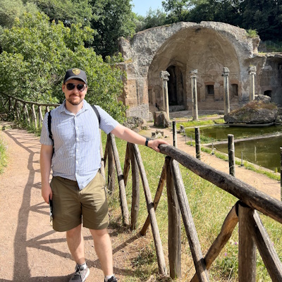 Picture of Professor Kevin Feeney in a light blue short sleeved shirt, olive green shorts, sunglasses, and a ball cap outside Hadrian's Villa.