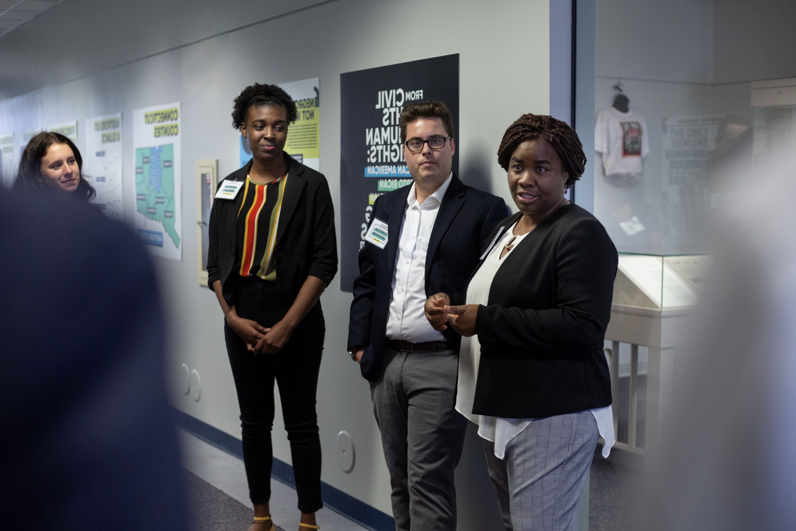 Four people stand in front of a display board discussing a topic with an audience. They are wearing business casual attire and appear to be part of an organized event or presentation.