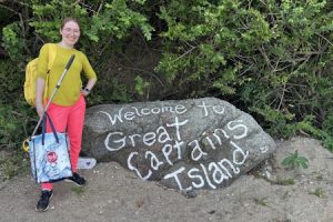 Kaitlyn Hiis LaRose, smiling, holding a shopping bag and a trash picker next to a large gray boulder with the words "Welcome to Great Captains Island" painted on it in white.